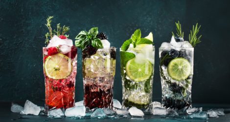 Cocktails drinks. Classic alcoholic long drink or mocktail highballs with berries, lime, herbs and ice on blue background