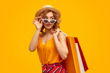 Trendy young woman with paper bags adjusting sunglasses and looking away over shoulder during shopping against bright yellow background