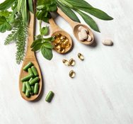 Natural vitamins, plant based supplements and green leaves on white wooden background, top view.