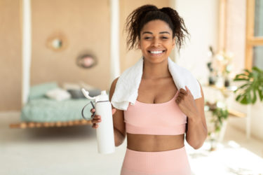 Portrait of fit black woman in sportswear holding shaker with whey protein or fresh water, working out at home.
