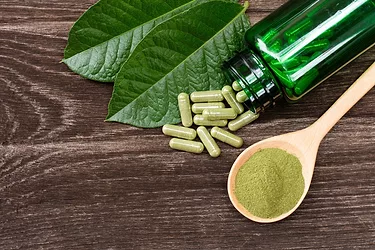 Kratom (Mitragyna speciosa) capsules with kratom powder and green leaves on wooden table background.