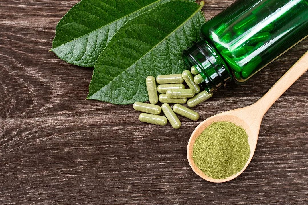 Kratom (Mitragyna speciosa) capsules with kratom powder and green leaves on wooden table background.