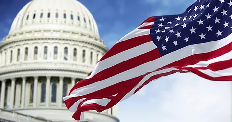 The national flag of the United States waving in the wind with the American US Capitol blurred in the background.