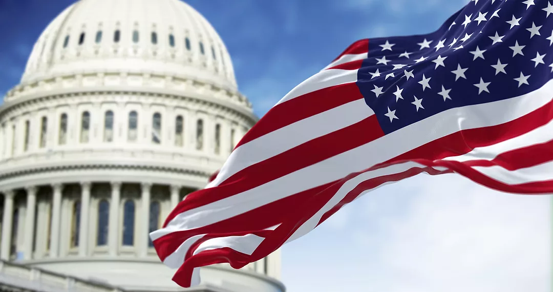 The national flag of the United States waving in the wind with the American US Capitol blurred in the background.